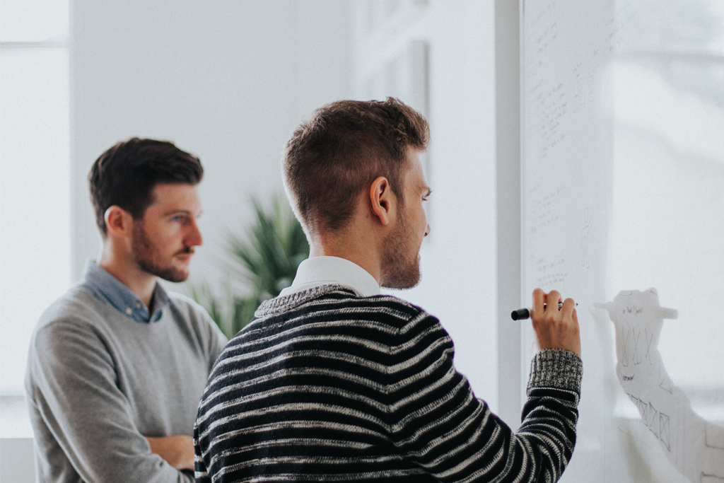 Zwei Männer, die im Coworking Space gemeinsam brainstormen und Notizen auf einem Whiteboard notieren